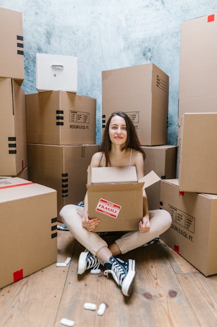 A young woman with long dark hair and a neutral expression is seated on a wooden floor inside a room surrounded by numerous cardboard boxes of various sizes, some sealed with packing tape and one mark