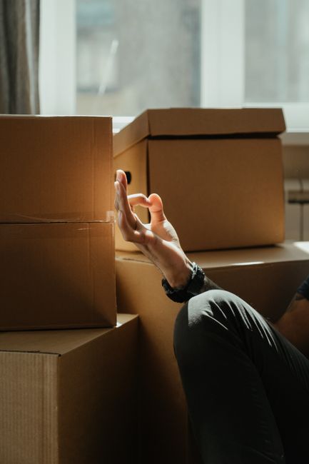 A young woman with long dark hair and a neutral expression is seated on a wooden floor inside a room surrounded by numerous cardboard boxes of various sizes, some sealed with packing tape and one mark